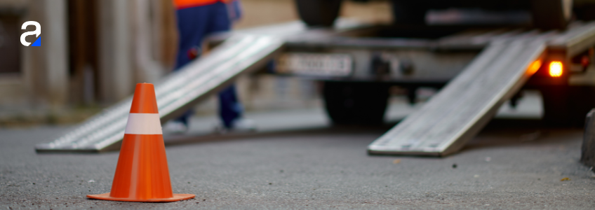 Roadside safety image with tow truck operator in high-visibility orange vest loading a vehicle onto a tow truck in the background with a safety cone in the foreground